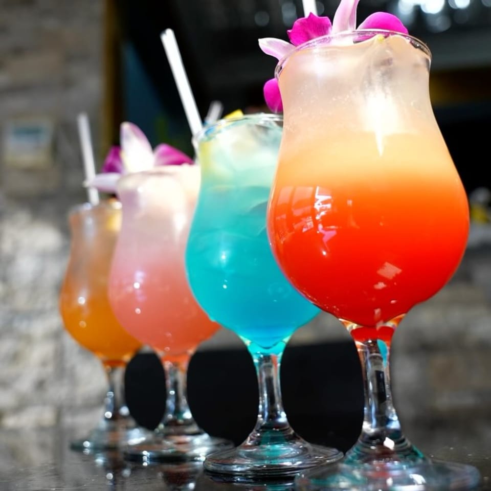 Four vibrant, colorful cocktails in tall glasses with straws and orchid garnishes on a dark surface.