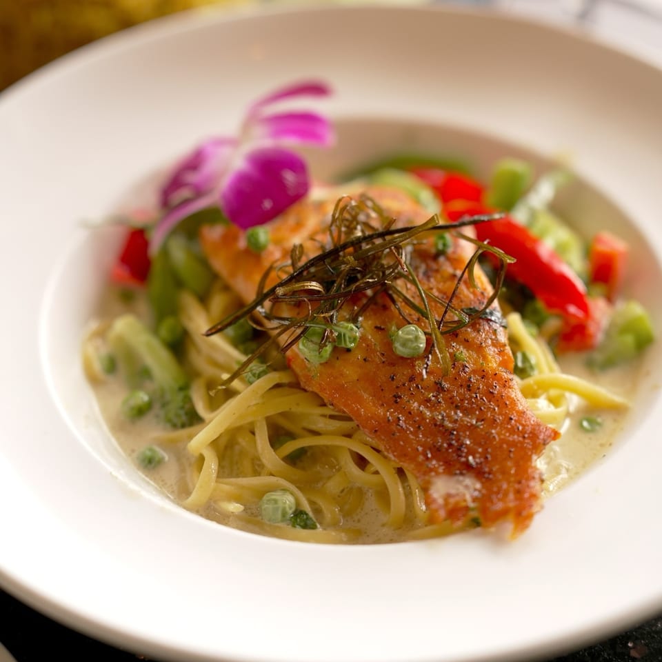 Pan-seared salmon over noodles with green curry, vegetables, and an orchid garnish.