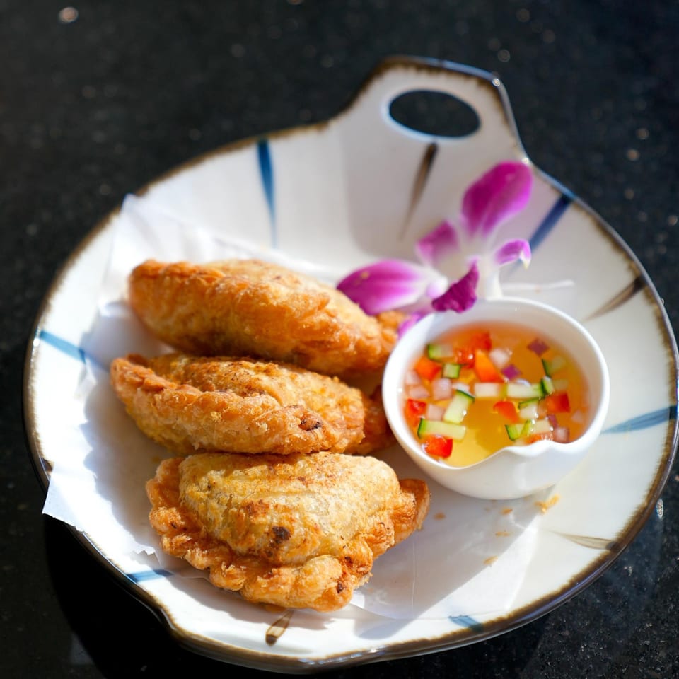 Golden-brown Thai curry puffs with a side of sweet chili dipping sauce and an orchid garnish.