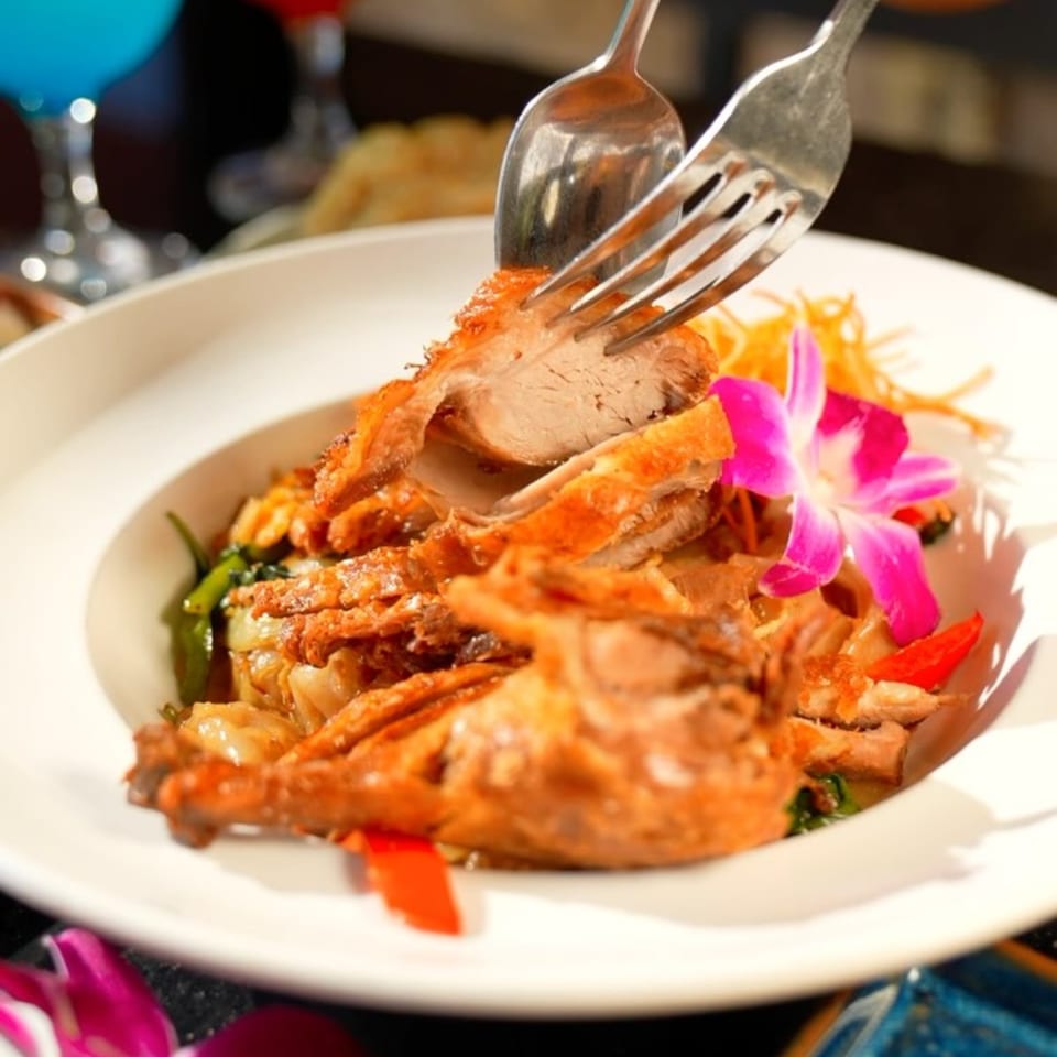 Crispy duck with noodles, vegetables, and a pink orchid garnish, being served with a fork and spoon.