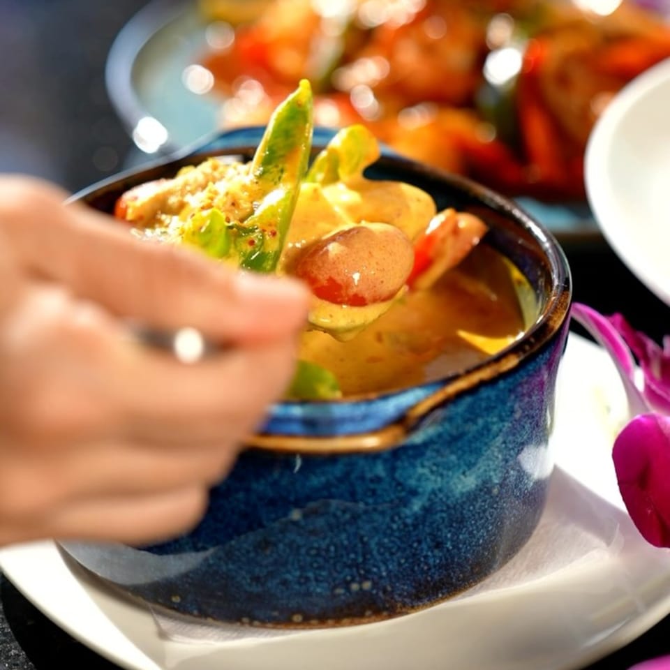 A hand spoons yellow curry with vegetables and potatoes from a blue bowl at Maya Thai Eatery.