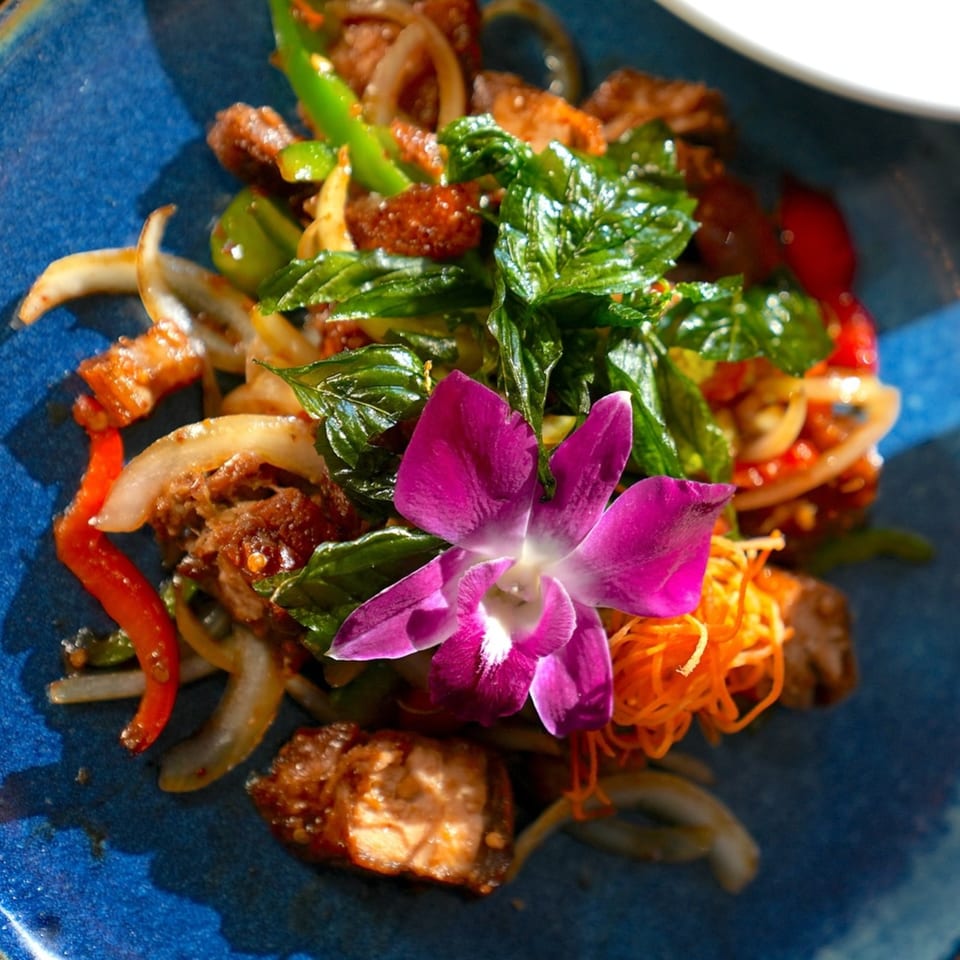 Close-up of a Thai dish with meat, vegetables, and a purple orchid garnish on a blue plate.